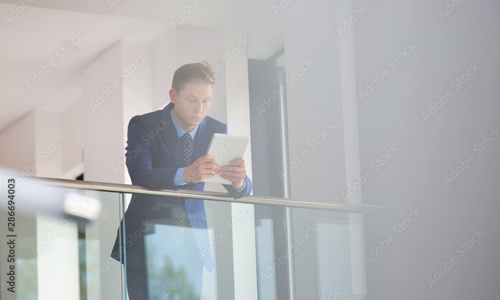 Young businessman using digital tablet while leaning on railing at office