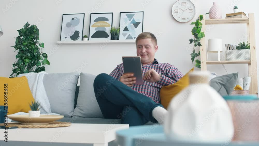 Man seat on sofa in living room and use tablet. Online shopping or social network surface. Happy young boy relaxing in home and stay online. 4k. Man seat on sofa in living room and use tablet. Online shopping or social network surface. Happy young boy relaxing in home and stay online. 4k.