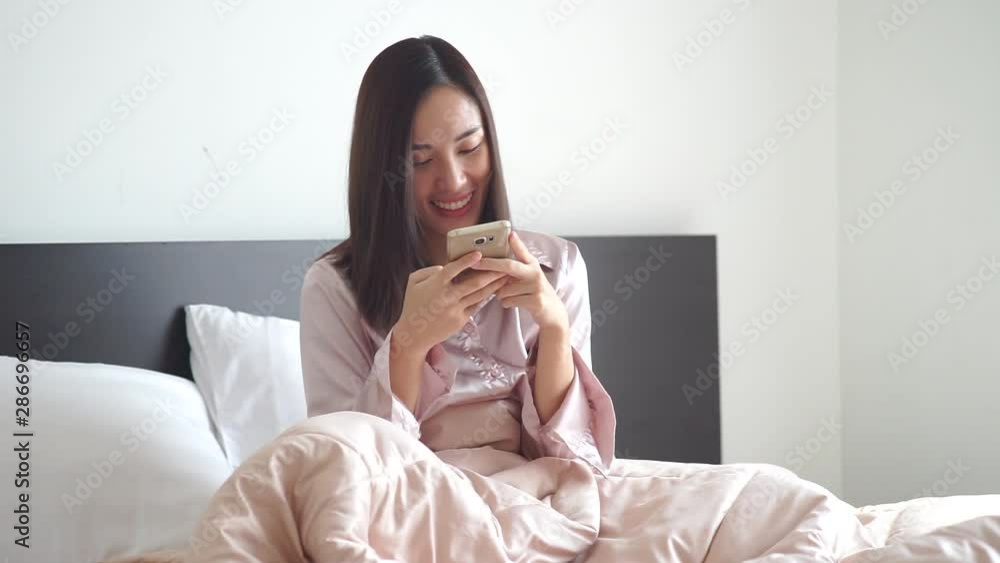 Attractive Asian woman wearing pajamas play game or search for products by phone on bed in the morning at her room Attractive Asian woman wearing pajamas play game or search for products by phone on bed in the morning at her room