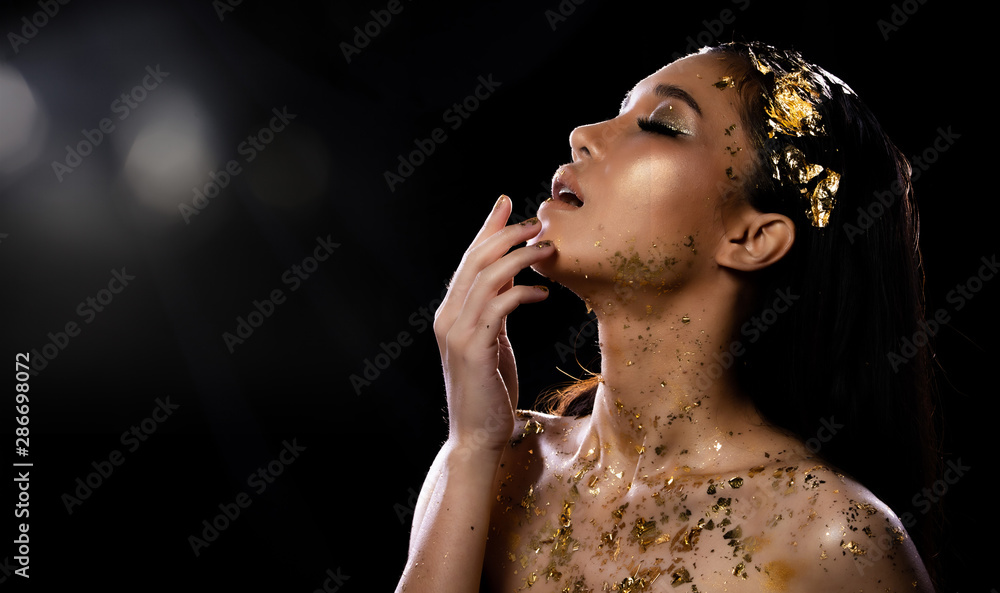 Obraz premium Fashion Young Asian Woman eyes brown black straight hair beautiful make up fashion decorate with Golden Foil or Gold leaf all over neck and shoulder. Studio Lighting dark Background star holes smoke