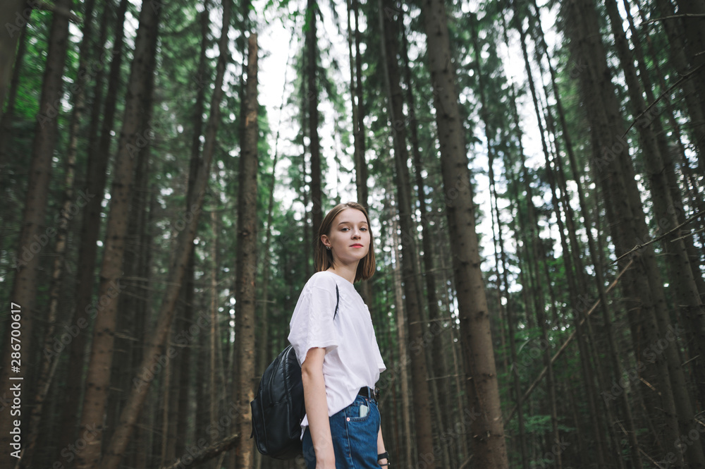 Portrait of a smiling female hiker with a backpack standing in the mountain coniferous forest looking at camera. Cheerful tourist girl standing among the fir trees on the woodland background. Portrait of a smiling female hiker with a backpack standing in the mountain coniferous forest looking at camera. Cheerful tourist girl standing among the fir trees on the woodland background.