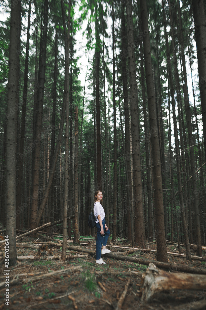 Vertical shot of caucasian tourist girl in white T-shirt walking in mountain fir forest feeling relaxed and calm. Attractive female hiker looking at camera standing in the middle of coniferous woods. Vertical shot of caucasian tourist girl in white T-shirt walking in mountain fir forest feeling relaxed and calm. Attractive female hiker looking at camera standing in the middle of coniferous woods.