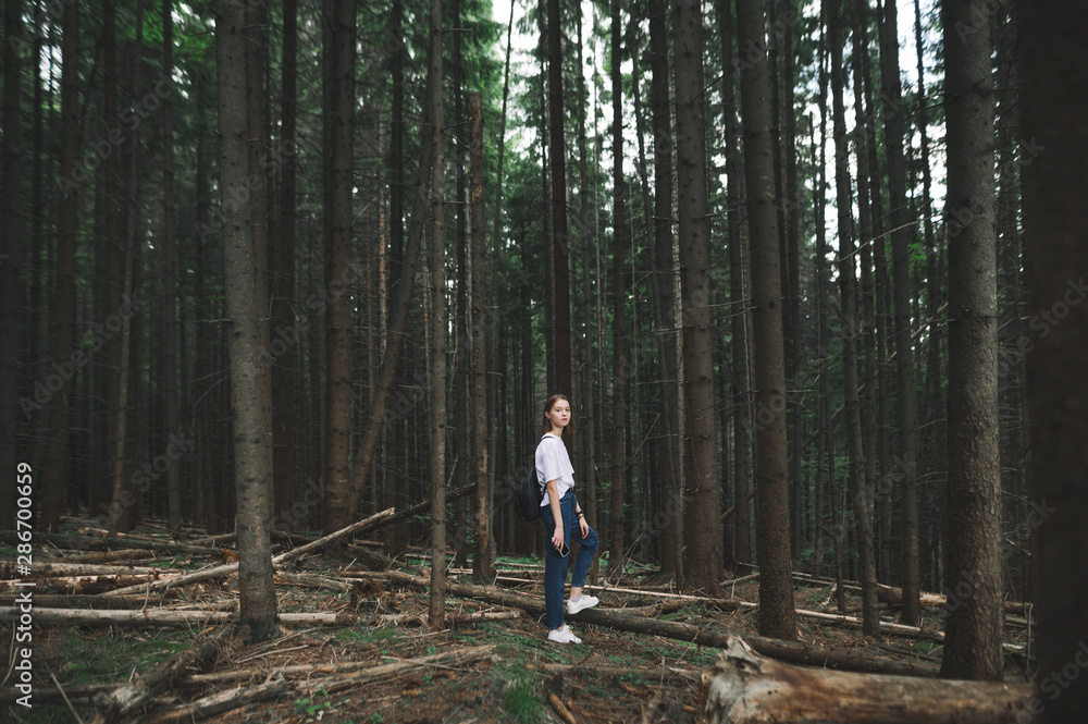 Young caucasian tourist girl in white T-shirt walking in the mountain fir forest feeling relaxed and calm. Attractive female hiker looking at camera standing in the middle of the coniferous woods.