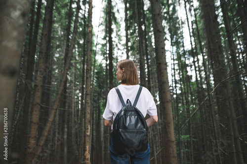 Back view on a female hiker with a backpack standing in the mountain coniferous forest looking aside. Tourist girl standing among the fir trees on the woodland background.