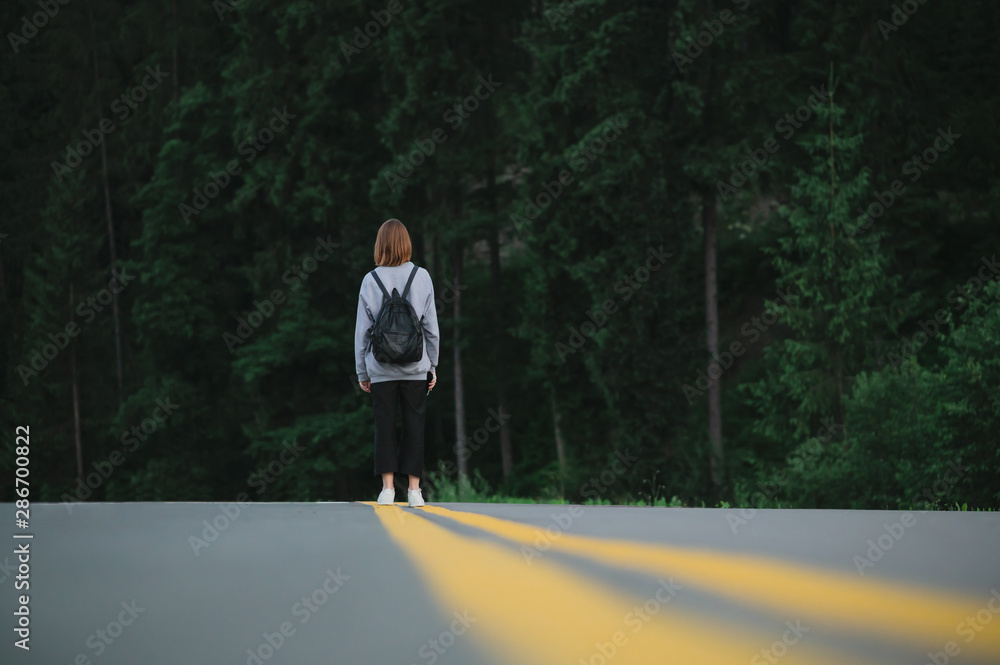 Obraz premium Horizontal shot of female traveler walking down the road at evening mountain forest background. Tourist girl with a backpack in the middle of the asphalt road near the woods.