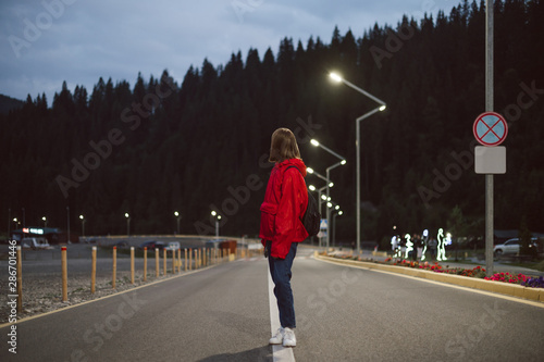 Back view of young female traveler walking down the asphalt road between the mountain forest in the evening. Tourist girl in a red raincoat strolling near the mountain fir forest in the street lights.