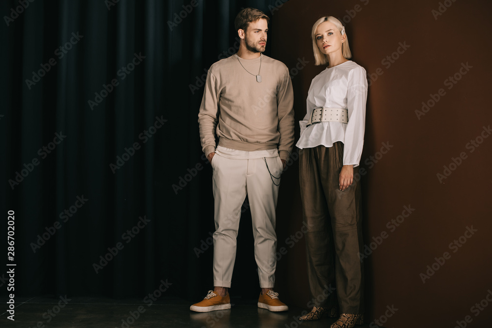 full length view of stylish man with hands in pockets and blonde woman in blouse standing near curtain - obrazy, fototapety, plakaty 