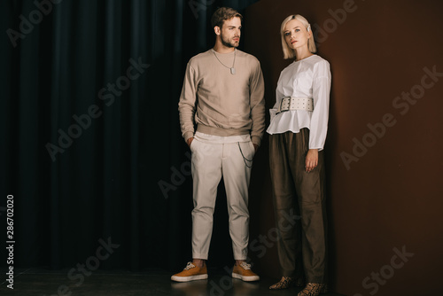 full length view of stylish man with hands in pockets and blonde woman in blouse standing near curtain