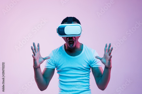 Young african-american man's playing in VR-glasses in neon light on gradient background. Male portrait. Concept of human emotions, facial expression, modern gadgets and technologies. Touches something