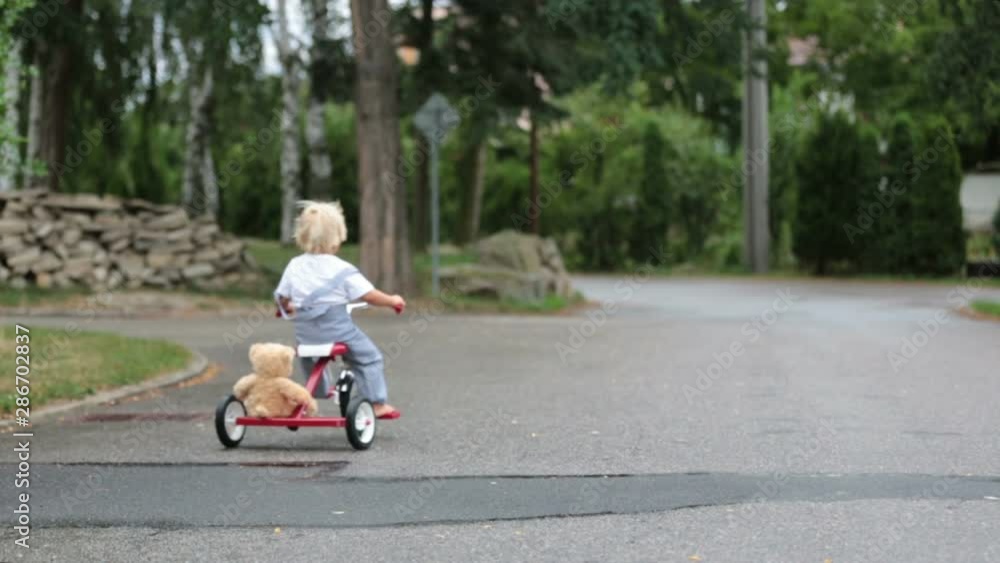 Cute little boy, with teddy bear toy, riding tricycle on the street in the rain, barefeet, summertime Cute little boy, with teddy bear toy, riding tricycle on the street in the rain, barefeet, summertime