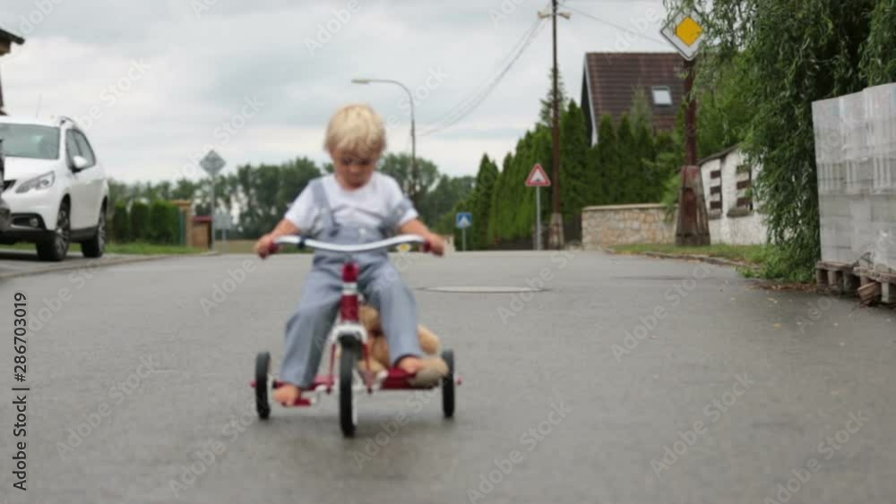 Cute little boy, with teddy bear toy, riding tricycle on the street in the rain, barefeet, summertime Cute little boy, with teddy bear toy, riding tricycle on the street in the rain, barefeet, summertime
