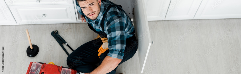 panoramic shot of bearded handyman sitting on floor near toolbox
