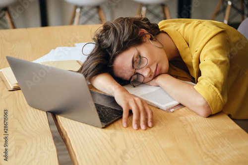 Young tired woman in eyeglasses asleep on desk with laptop and documents under head at workplace photo