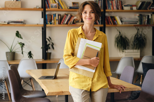 Young smiling woman in yellow shirt leaning on desk with notepad and papers in hand while happily looking in camera in modern office