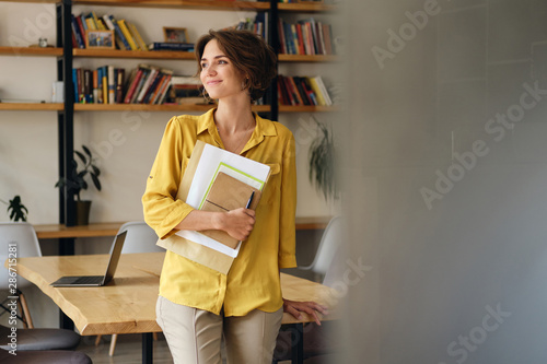 Young attractive woman in yellow shirt leaning on desk with notepad and papers in hand while dreamily looking aside in modern office