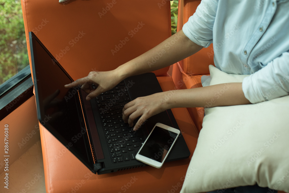 Naklejka premium Women sit in a coffee shop on orange sofa using a computer laptop and mobile phone. To contact business or to talk to a friend or shopping online.