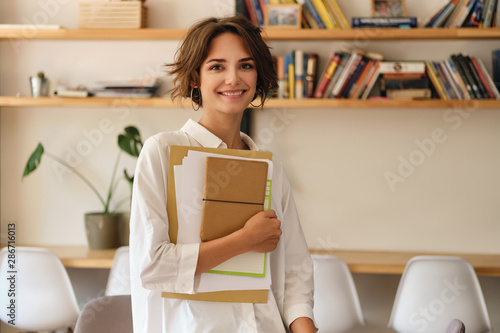 Young attractive smiling woman joyfully looking in camera with papers and notepad in office