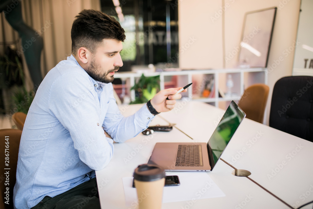 Fototapeta premium Young businessman working in office, sitting at desk, looking at laptop and pointed with pen on screen