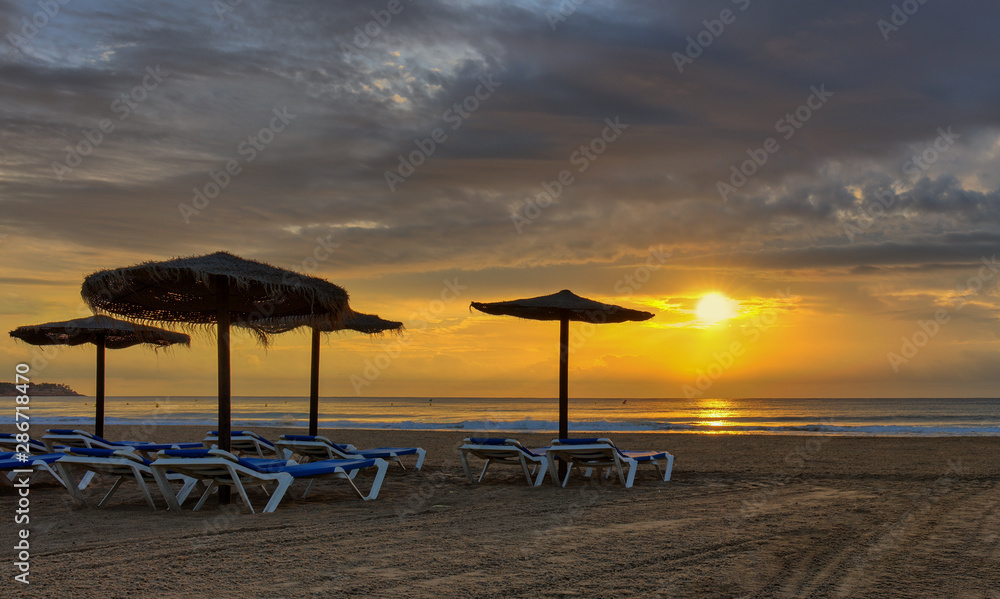 A dream beach with umbrellas made of straw and blue beach chairs in the early morning at sunrise on the Mediterranean sea. Beautiful clouds adorn the sky.