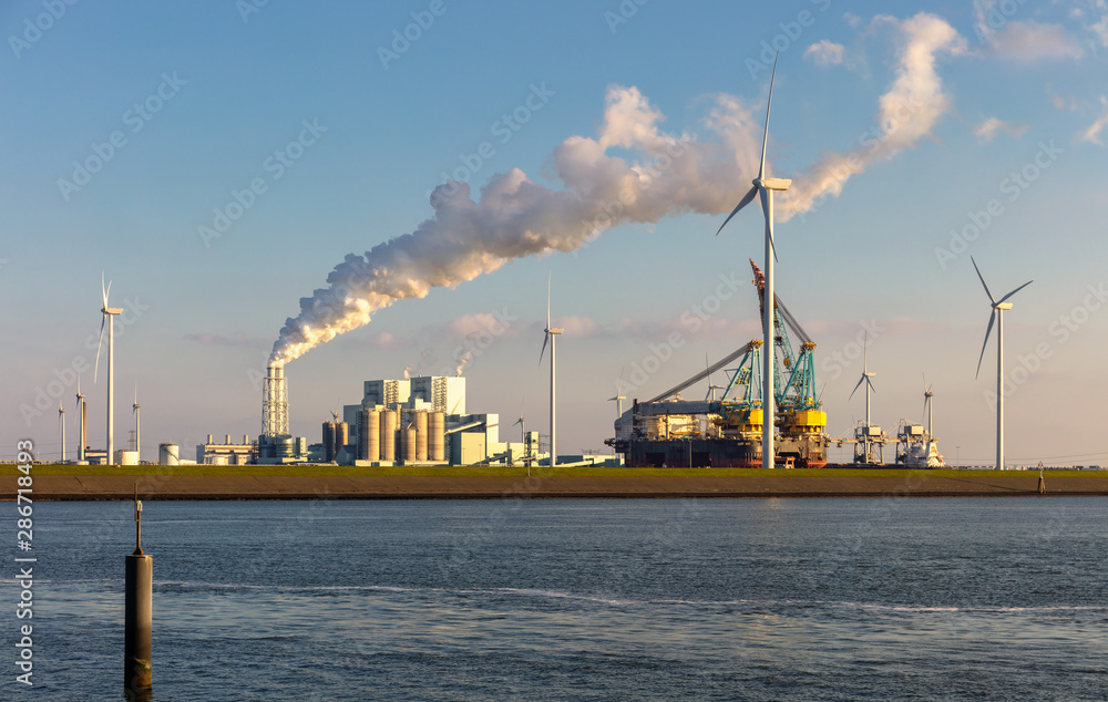 Obraz premium Widok na przemysłową przystań Eemshaven w Holandii z dymiącymi kominami. Na pierwszym planie basen portowy. Statek, wiele turbin wiatrowych i błękitne niebo.