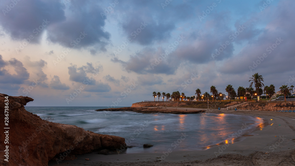 A beautiful bay with sandy beach and palm trees at early morning before sunrise near the Spanish town of Torrevieja. The lanterns are still lit and some beautiful clouds are in the sky.