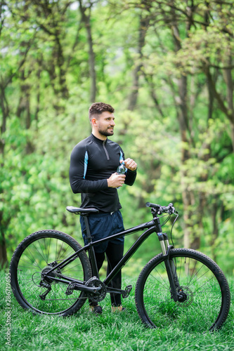 Young man in track-suit with bicycle drinking water at summer park
