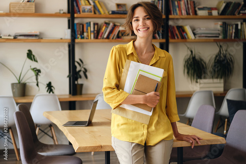 Young cheerful woman in yellow shirt leaning on desk with notepad and papers in hand while joyfully looking in camera in modern office