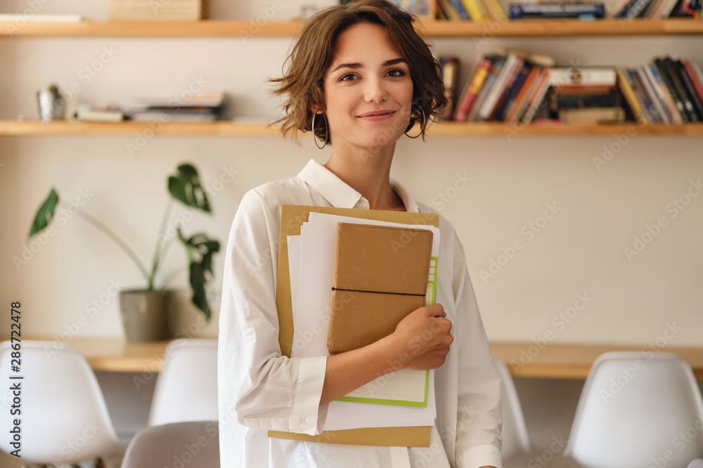 Young beautiful smiling woman dreamily looking in camera with papers and notepad in office - obrazy, fototapety, plakaty 