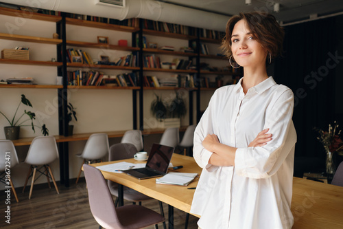 Young attractive woman in white shirt dreamily looking in camera with desk on background in office photo