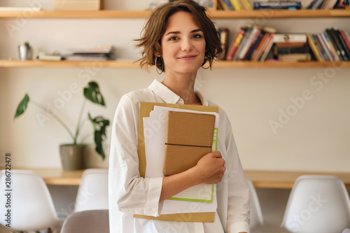 Young beautiful smiling woman dreamily looking in camera with papers and notepad in office