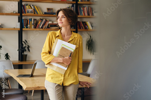 Young beautiful woman in yellow shirt leaning on desk with notepad and papers in hand while thoughtfully looking aside in modern office