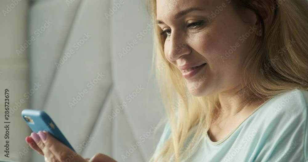 Adult blonde woman using mobile phone, at home. Adult beautiful woman typing message on a smartphone. Happy smiling woman uses cell phone lying on bed. Joyful woman holds smart phone in hands. Adult blonde woman using mobile phone, at home. Adult beautiful woman typing message on a smartphone. Happy smiling woman uses cell phone lying on bed. Joyful woman holds smart phone in hands.