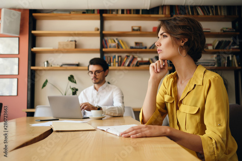 Side view of young woman sitting at the table with notepad thoughtfully working with colleague on background in office