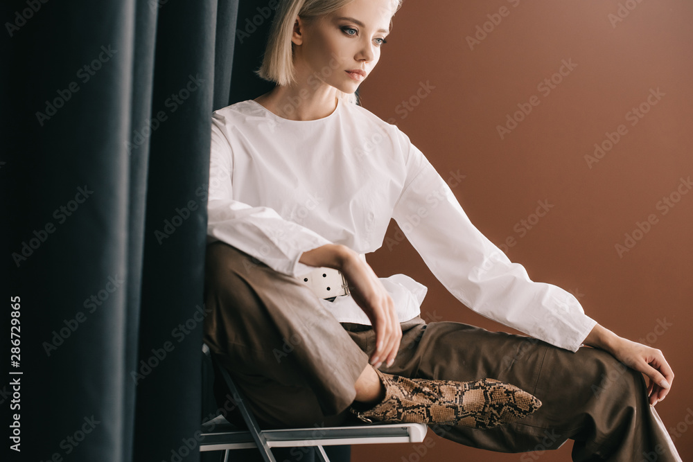 Stylish blonde woman in white blouse sitting on chair near curtain on brown - obrazy, fototapety, plakaty 