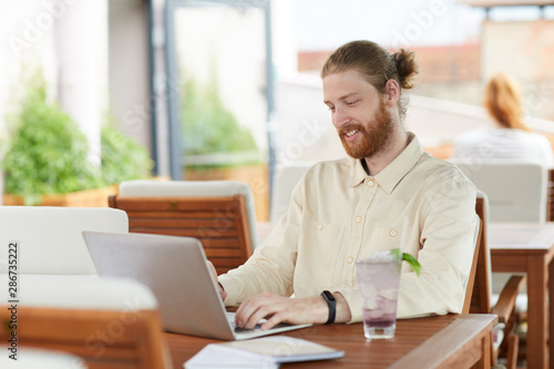 Young bearded man communicating online on his laptop and smiling while sitting at the table in cafe