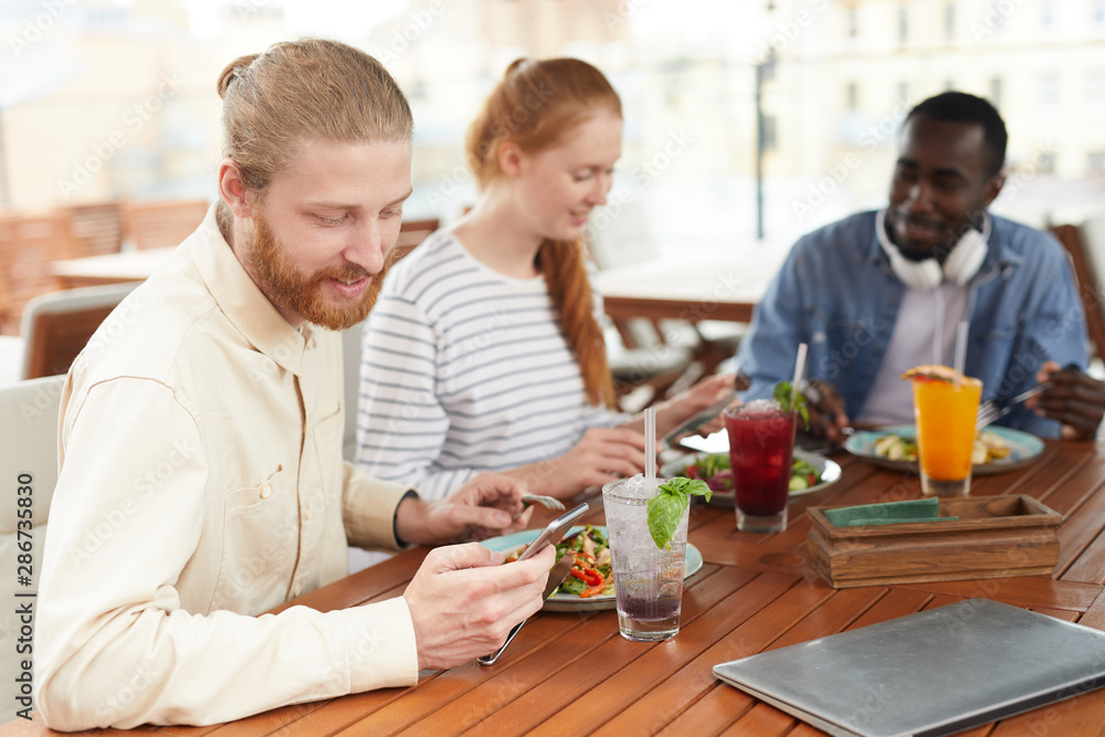 Young bearded man using mobile phone and smiling while have a lunch together with his friends in cafe Young bearded man using mobile phone and smiling while have a lunch together with his friends in cafe