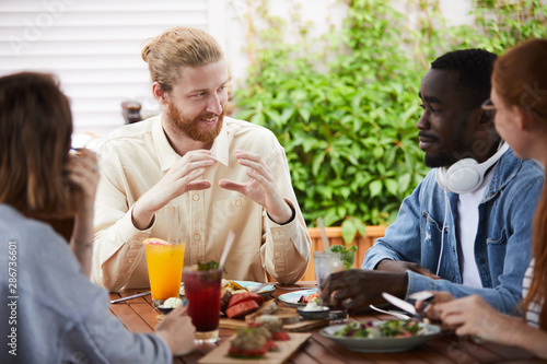 Young bearded man sitting at the table gesturing and explaining something to young people during their lunch in cafe