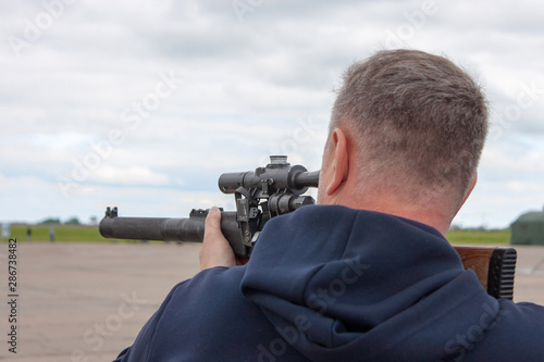 A short-haired man in a blue sweater with a hood is aiming from an army gun. Back view