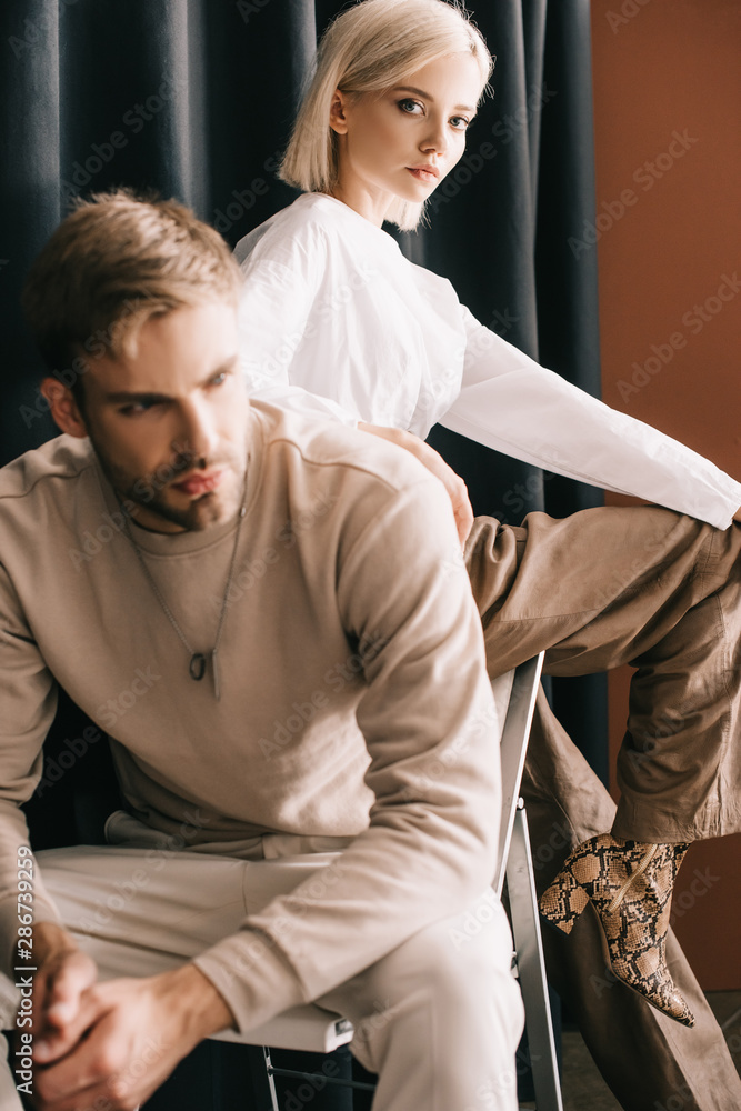 blonde woman in white blouse and bearded man sitting on chair near curtain - obrazy, fototapety, plakaty 