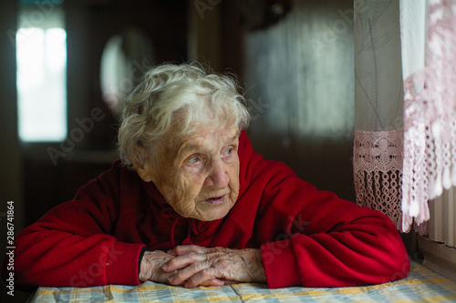 An old russian woman sits in a house near the window.