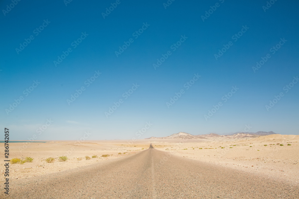 Road in desert stretching into the distance. Mountains on the horizon. Clear blue sky, scorching sun. Nobody around Road in desert stretching into the distance. Mountains on the horizon. Clear blue sky, scorching sun. Nobody around