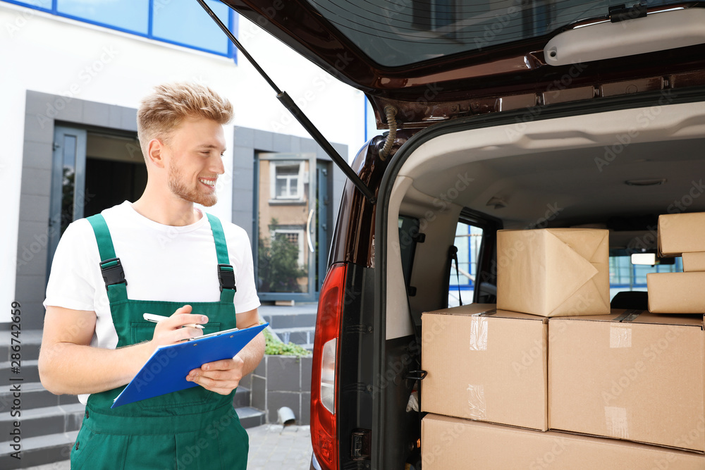 Young courier holding clipboard near delivery van with parcels outdoors Young courier holding clipboard near delivery van with parcels outdoors