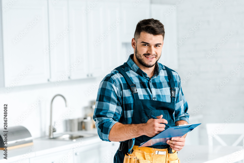 happy bearded workman holding clipboard and looking at camera in kitchen happy bearded workman holding clipboard and looking at camera in kitchen