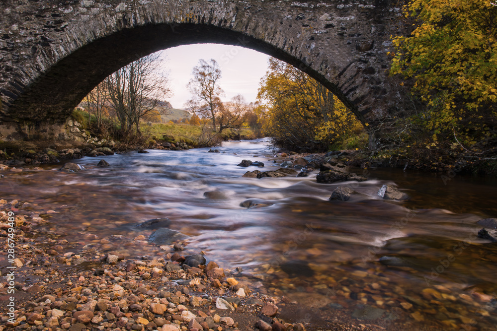 Long exposure photograph under the old military bridge along the 18th century military road in Perthshire in Scotland. Long exposure photograph of creek. Autumnal scene in the background.