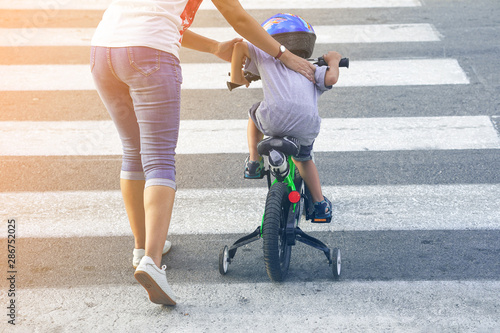 Mother goes pedestrian crossing with childr on bicycle. A woman with son crossing the road in the city. Back view.