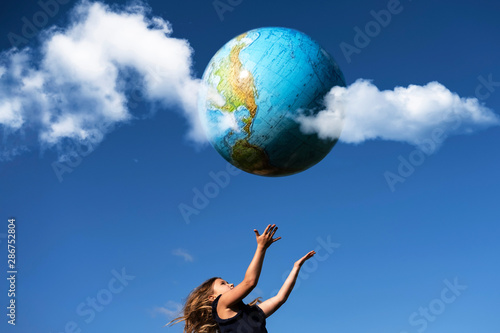 Young girl catches planet Earth. Happy child with globe in raised hands on blue sky background. Environment and ecology protection concept. World Earth Day greeting card, postcard idea