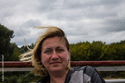 White blonde woman 42 years old with short hair flying in the wind on Board the ship against the river and the banks with green trees and cloudy sky