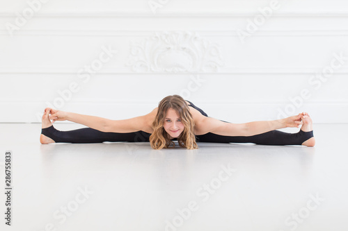 Blond woman in sport outfit doing splits on white bare floor