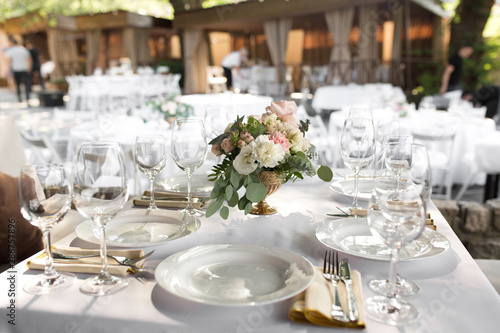 Wedding table setting decorated with fresh flowers in a brass vase. Wedding floristry. Banquet table for guests outdoors with a view of green nature. Bouquet with roses, eustoma and eucalyptus leaves