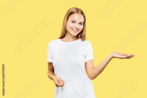 Caucasian young woman's half-length portrait on yellow studio background. Beautiful female model in white shirt. Concept of human emotions, facial expression. Showing and pointing something.